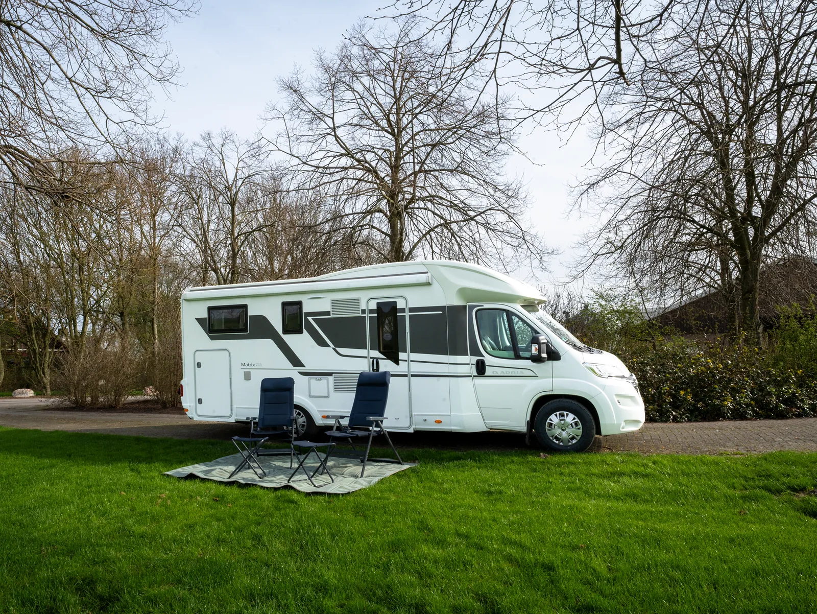 Adria Matrix 670 SL met uitgeklapte luifel, campingstoelen en tafel op een grasveld
